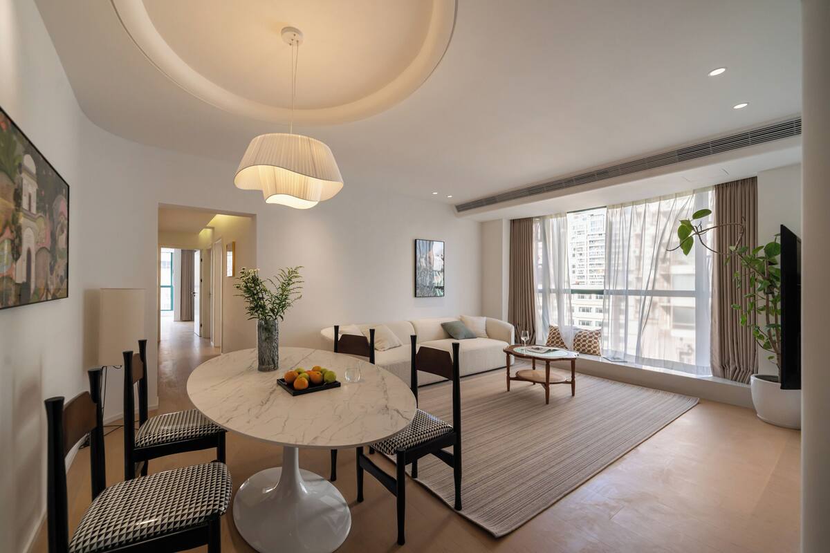 A modern living room with a round marble table, a sofa, and a coffee table. There is a large window with curtains, a potted plant, and a painting on the wall.
