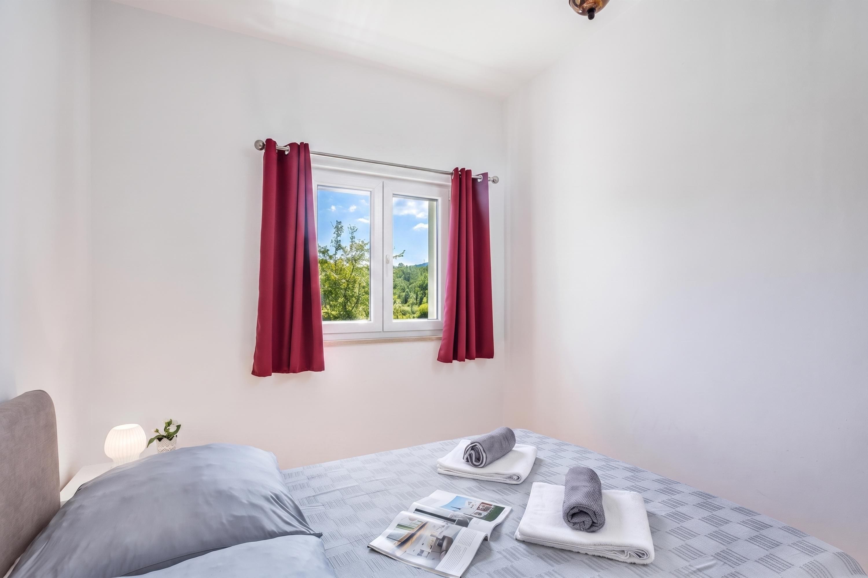 A bedroom with a bed, a window with curtains, and a view of greenery outside.
