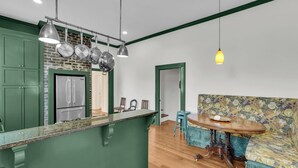 Una cocina con armarios verdes, una pared de ladrillo y ollas colgantes. Hay una mesa de comedor de madera, un sofá floral y una lámpara colgante.