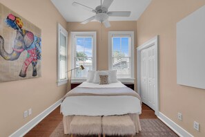 A bedroom with a bed, a ceiling fan, two windows with blinds, a door, and a wall-mounted artwork of an elephant.