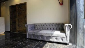 A grey tufted sofa in a lobby with a wooden door and a fire extinguisher on the wall. - Hotel Gorrion (Mumbai)