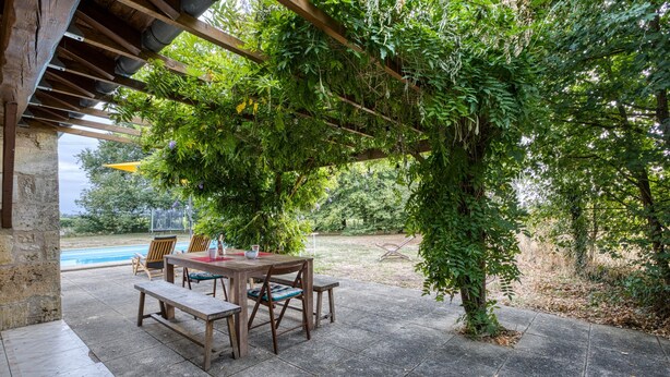 An outdoor patio with a wooden table and chairs, surrounded by lush greenery and a view of a swimming pool. - Holiday Home 'La Musardière' with Private Pool, Private Terrace and Wi-Fi (Champagnolles)