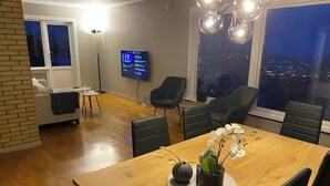 Ein Essbereich mit Holztisch, schwarzen Stühlen, einem Fernseher an der Wand und Blick auf die Stadt durch große Fenster.