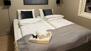 Ein ordentlich bezogenes Bett mit weißen Leinen, einer gemusterten Decke und einem gefalteten Handtuch. An der Wand über dem Bett hängt ein gerahmtes Bild von einer Berglandschaft.