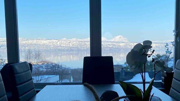 Ein Zimmer mit großem Fenster mit Blick auf einen See und eine verschneite Landschaft. Es steht ein Tisch mit einem Laptop, einem Topf mit einer Orchidee und einer Vase mit Blumen darauf.