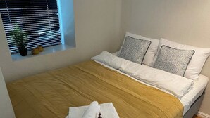A neatly made bed with a brown bedspread and white pillows, a window with blinds, and a small plant on the windowsill. - Central Narvik Apartment, Walk to Train & Bus (Narvik)