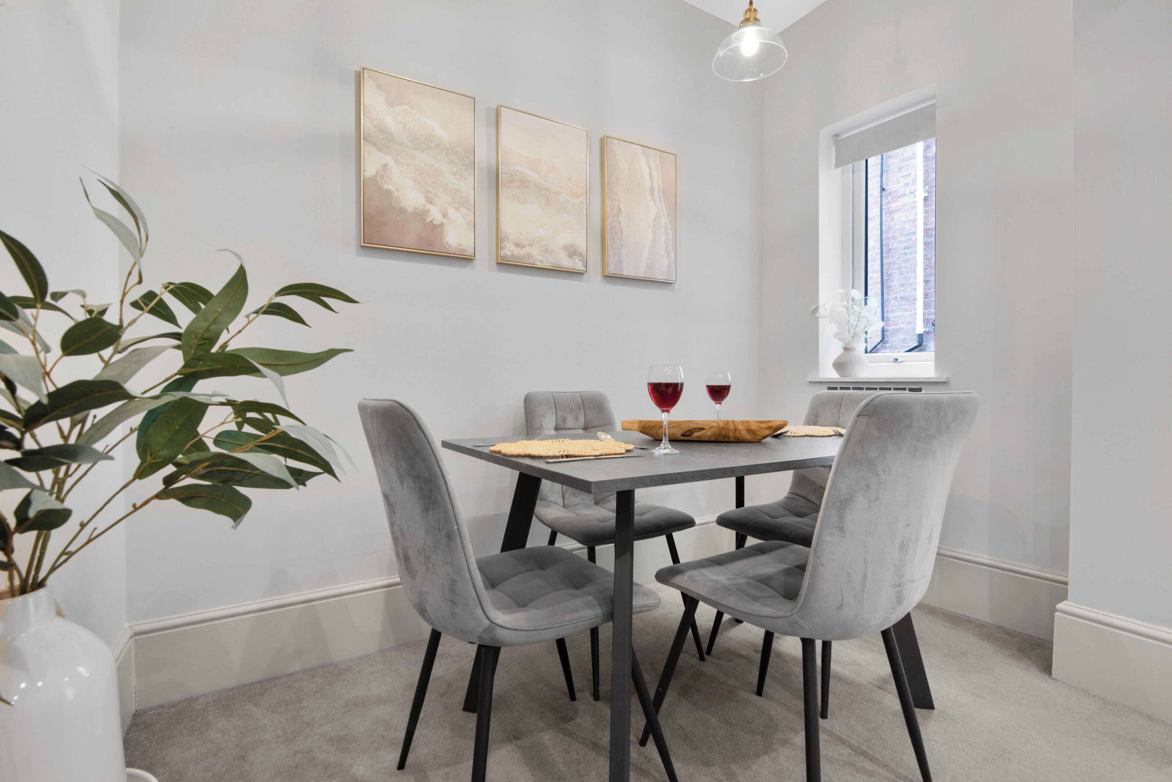 Un comedor con una mesa, cuatro sillas grises, un florero con hojas verdes y tres pinturas abstractas en la pared.