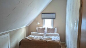 Une chambre avec un plafond en pente, un lit avec une literie blanche, une lampe de chevet et une fenêtre avec des stores.