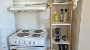 Une cuisine de taille réduite, avec un fourneau et un four blancs, ainsi qu’une étagère en bois où sont rangés des ustensiles de cuisine.