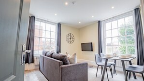 A modern living room with a grey sofa, a dining table with chairs, a flat-screen TV, and large windows with curtains. - WhiskAwayHomes Shaw Street Apartment 5 (Worcestershire)