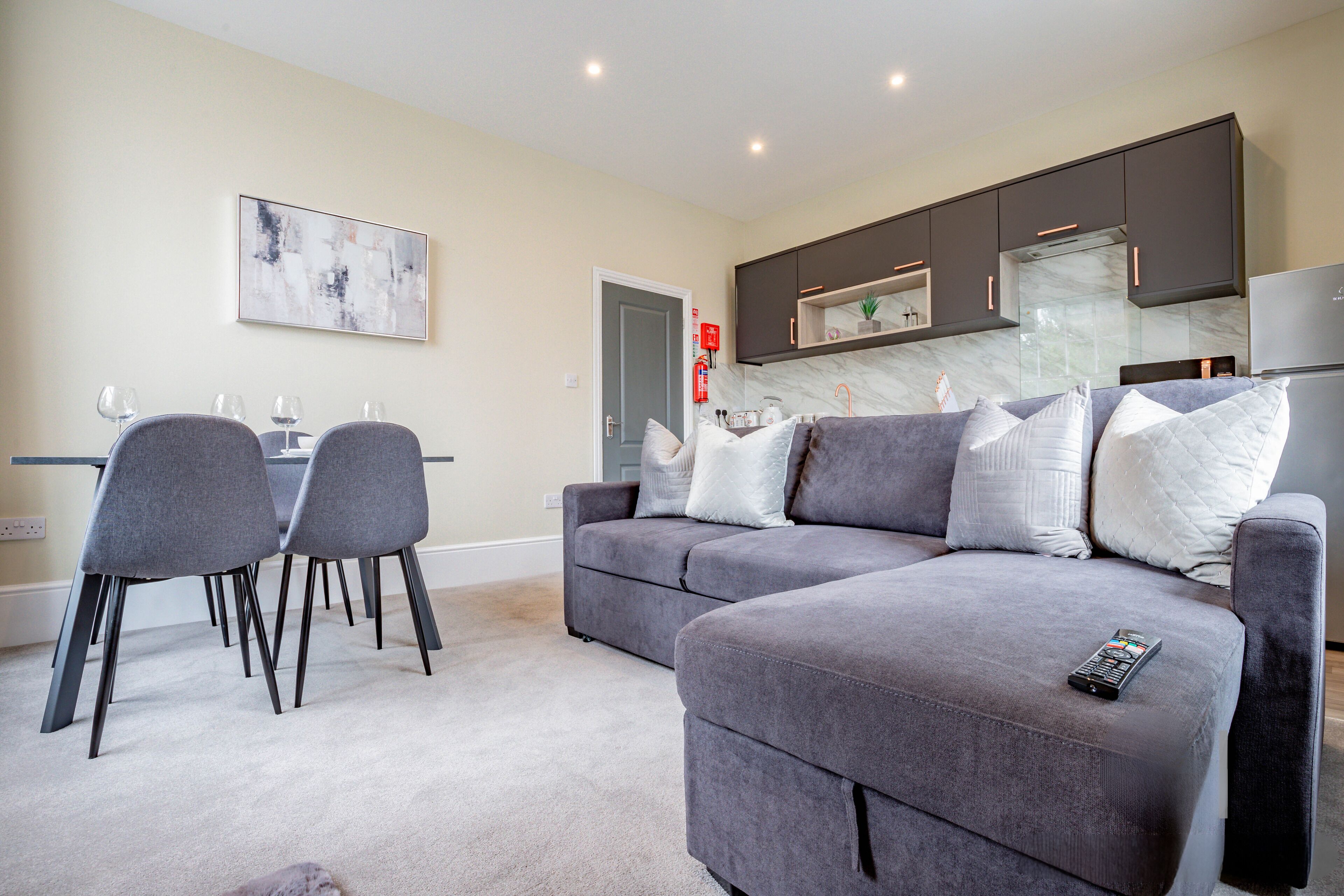 A modern living room with a grey sofa, a dining table with chairs, and a kitchen area with dark cabinets and a marble backsplash.