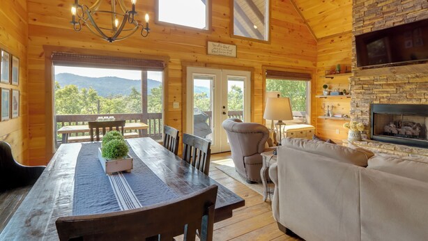 Interior de una cabaña de madera con una mesa de comedor, sillas, un sofá, una chimenea y vistas a las montañas a través de amplios ventanales.