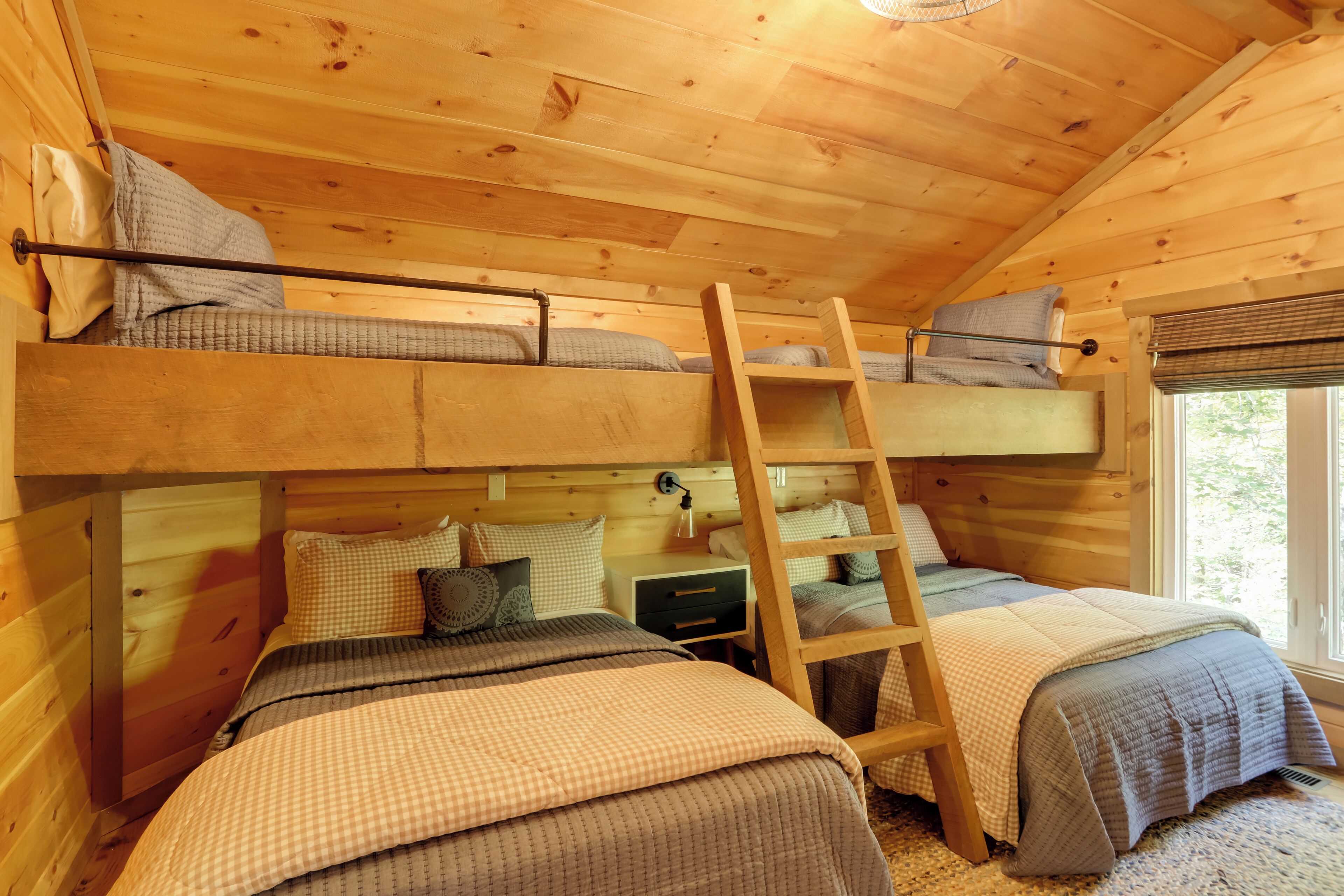 A cabin with two bunk beds, a ladder, a nightstand, and a window with a view of trees.