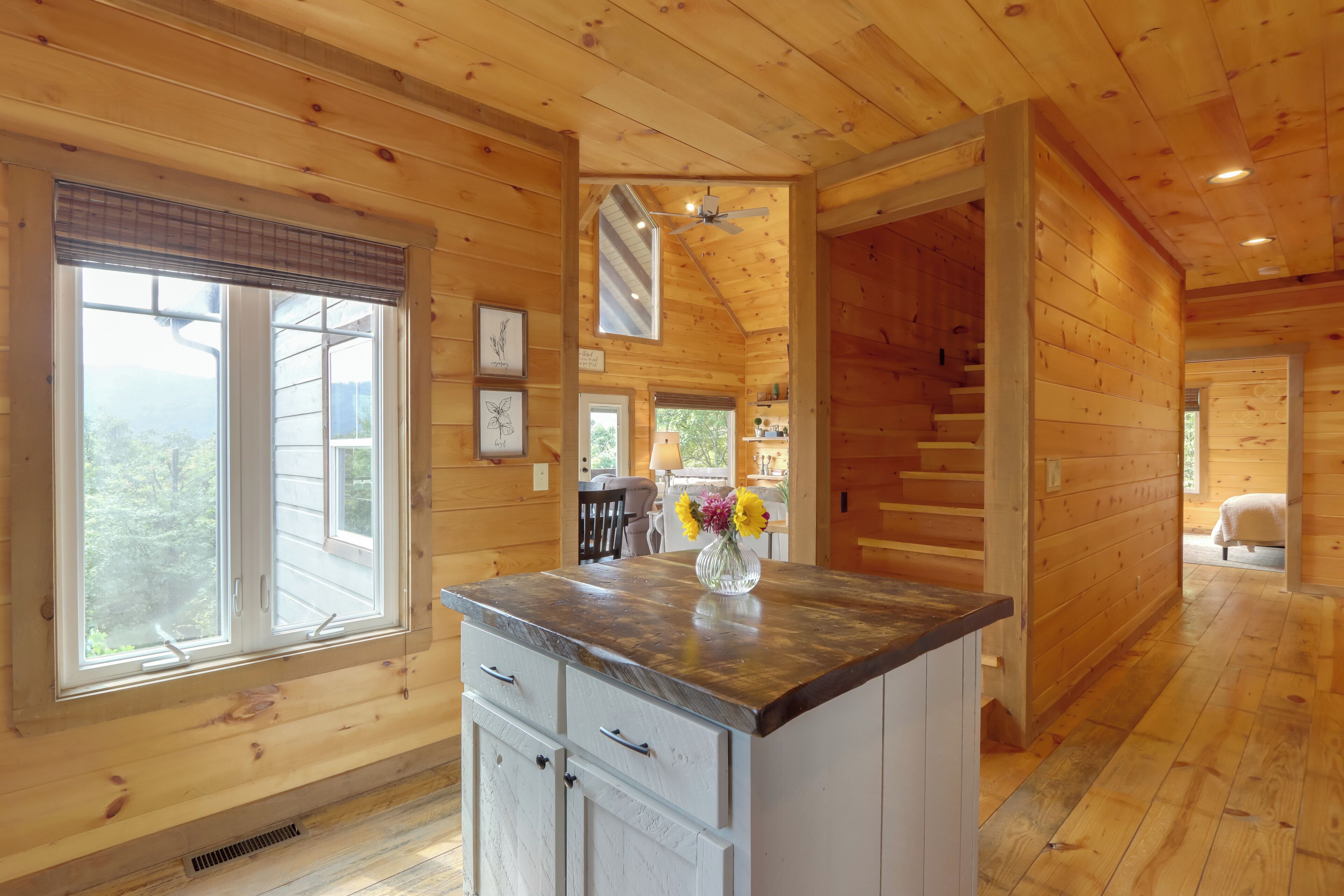 A wooden cabin interior with a kitchen area featuring a central island, a dining table, and a staircase leading to another level.