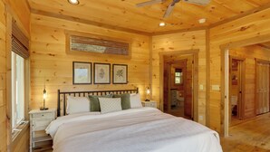 Un dormitorio de cabaña de madera con una cama, mesitas de noche, un ventilador de techo y cuadros en la pared.