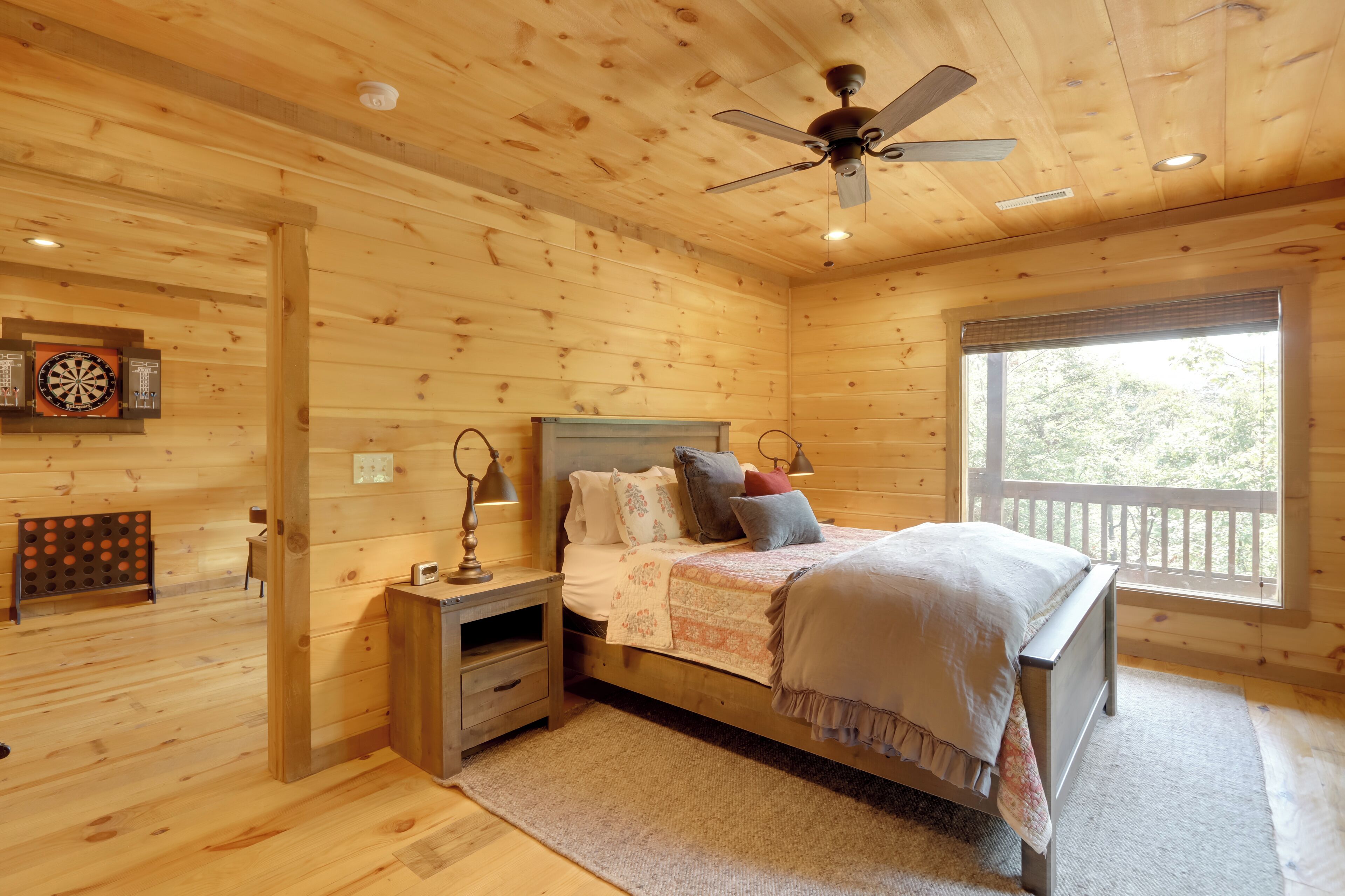 Un dormitorio de cabaña de madera con una cama, una mesita de noche, un ventilador de techo y una ventana con vistas a árboles.
