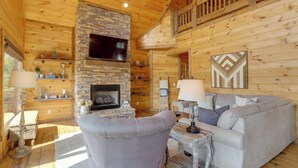 Interior acogedor de una cabaña de madera con chimenea de piedra, un televisor de pantalla plana montado encima y un sofá gris, además de un techo de madera.