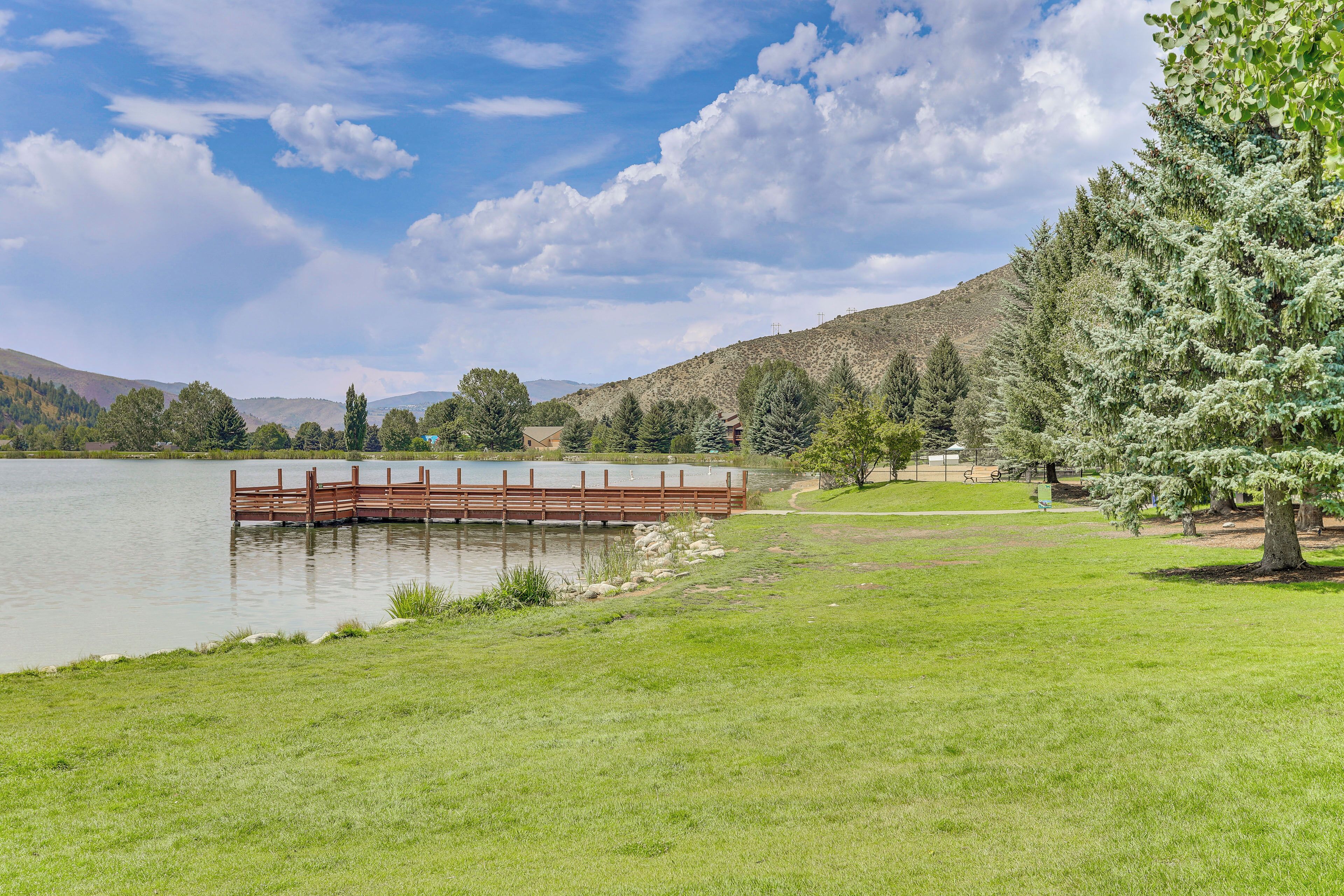 A serene lakeside setting with a wooden bridge over the water, grassy areas, and trees.