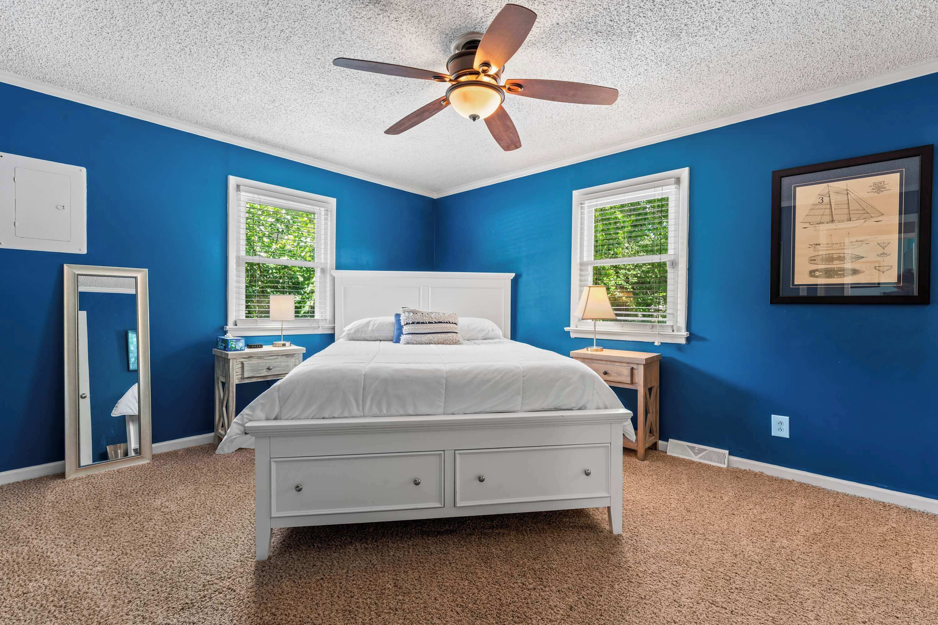Un dormitorio con una cama blanca, dos mesitas de noche, un ventilador de techo y dos ventanas con persianas.