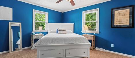 Un dormitorio con una cama blanca, dos mesitas de noche, un ventilador de techo y dos ventanas con persianas.