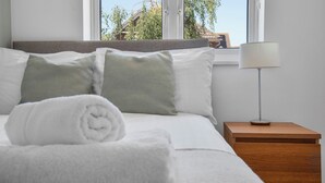 A neatly made bed with white linens and a rolled towel, a bedside table with a lamp, and a window with a view of trees and a building. - WhiskAwayHomes Pirie Avenue No. 2 (Worcestershire)