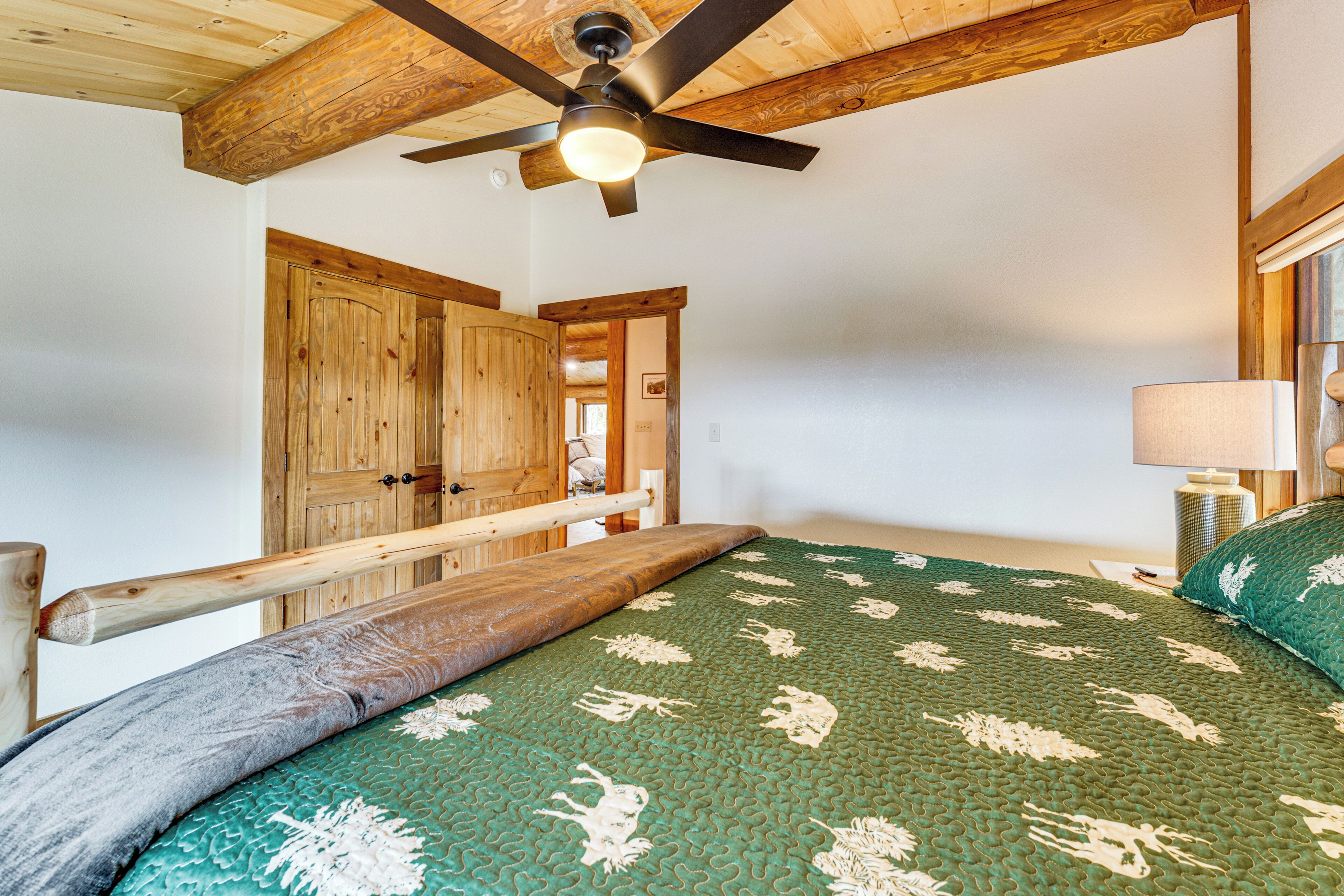 A bedroom with a wooden ceiling, a ceiling fan, a bed with a green patterned bedspread, and a wooden wardrobe.