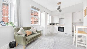 Un appartement moderne doté d’un espace de vie comprenant un canapé, une table à manger avec des chaises, une cuisine avec des meubles blancs et une grande fenêtre offrant une vue sur les immebles.