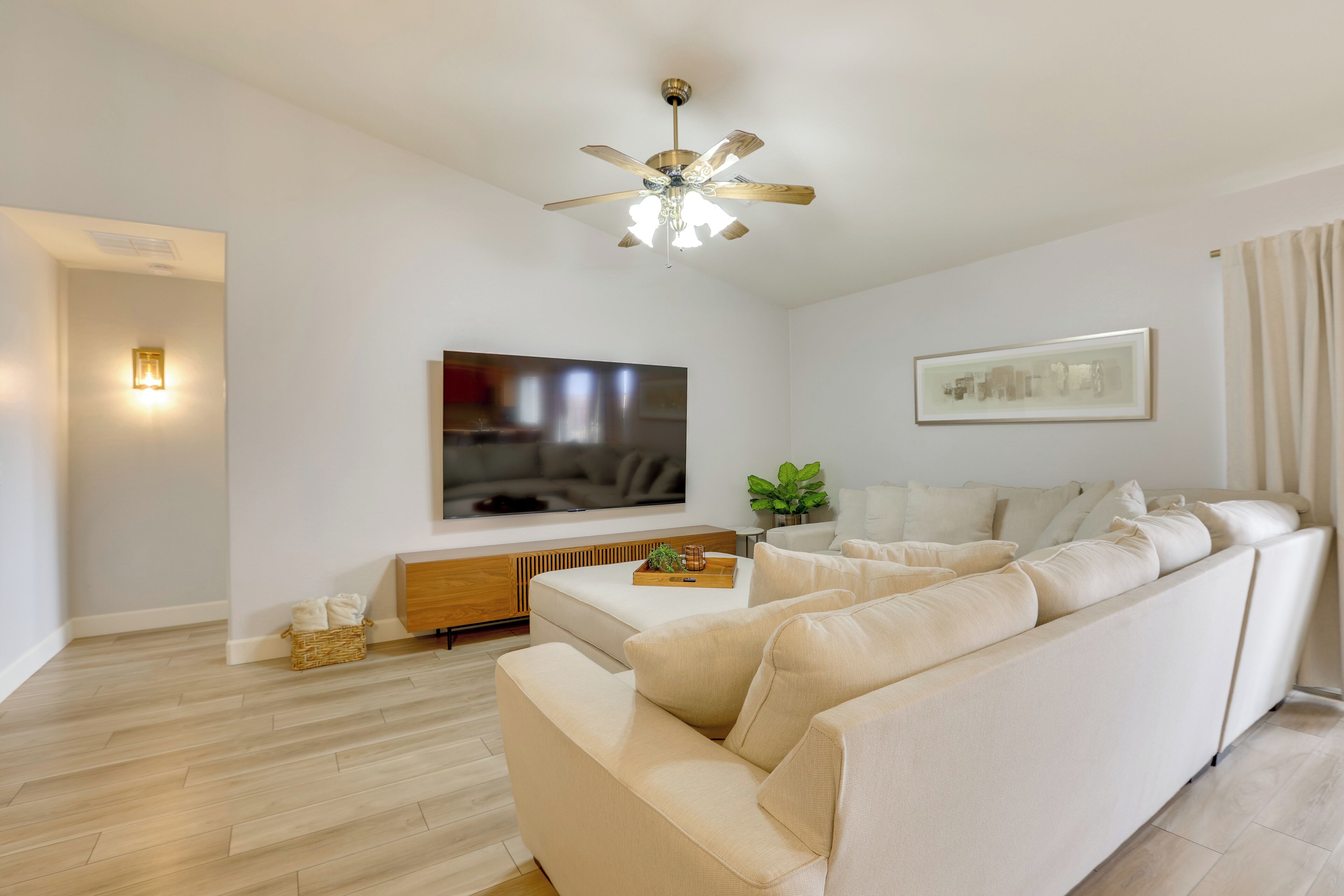 A modern living room with a large beige sofa, a flat-screen TV mounted on the wall, a ceiling fan, and a framed picture.