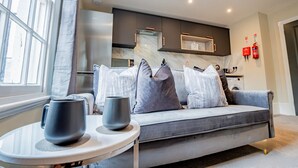 Een moderne woonkamer met een grijze bank, een ronde tafel met twee mokken, en een keuken op de achtergrond.