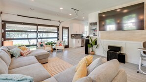 Un salon moderne avec un grand canapé d’angle, un téléviseur à écran plat fixé au mur et une cuisine en arrière-plan.