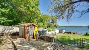 Une terrasse en bois menant à un petit chalet au bord d’un lac, avec une cour clôturée et une pile de bois de chauffage.