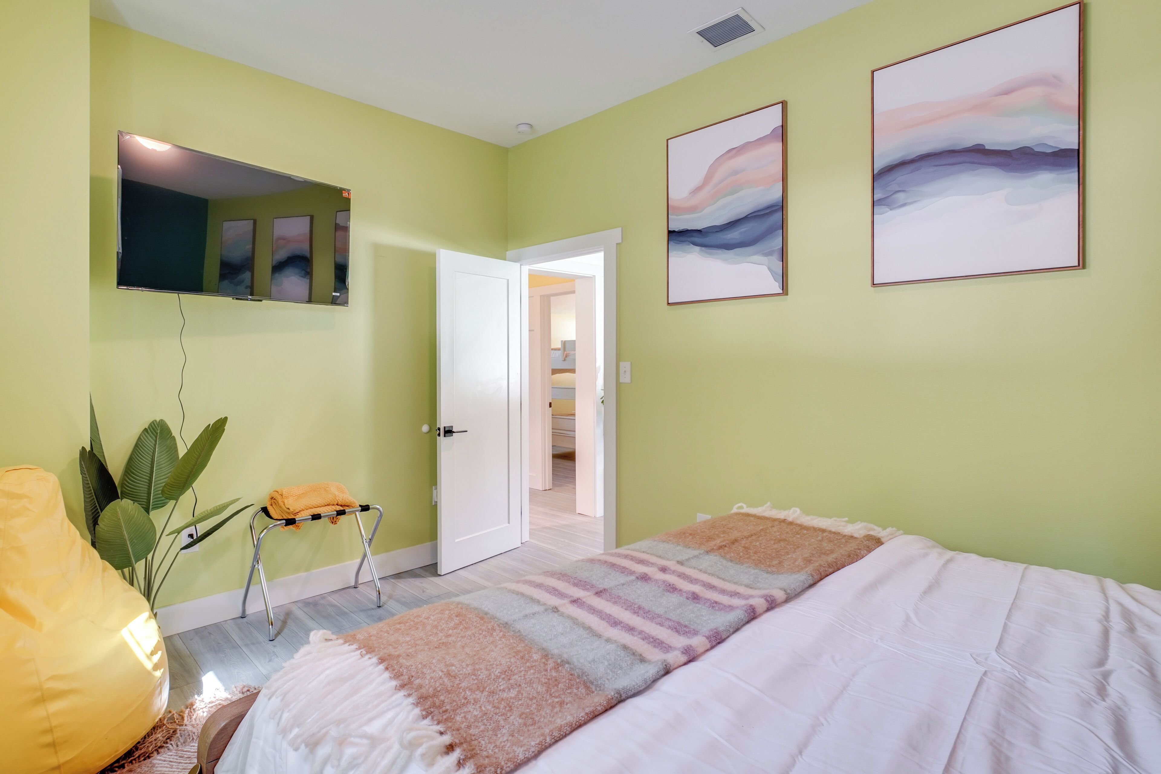 A bedroom with a bed, a small stool, a chair, and a plant.