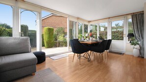 A spacious room with a grey sofa, a dining table with chairs, and large windows offering a view of the garden and neighboring buildings.