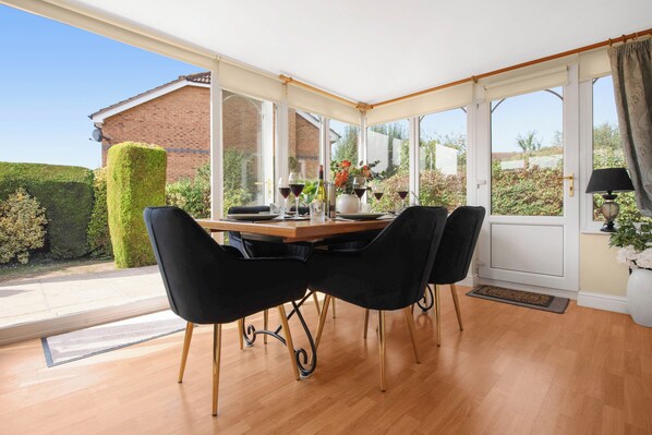 A conservatory with a wooden table and black chairs, large windows, and a view of a garden and house.