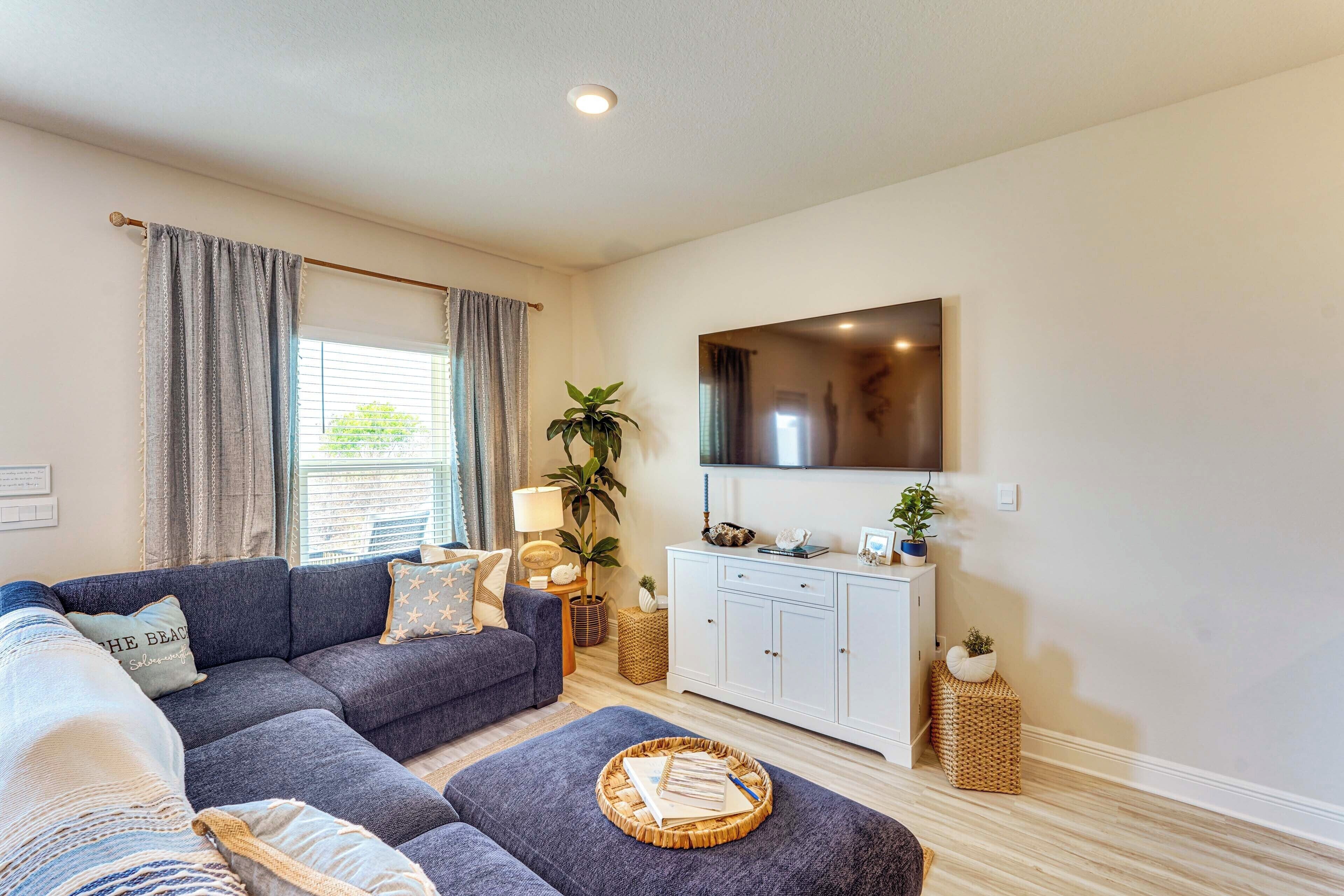 A living room with a sectional sofa, a flat-screen TV mounted on the wall, a white cabinet, and a large window with curtains.