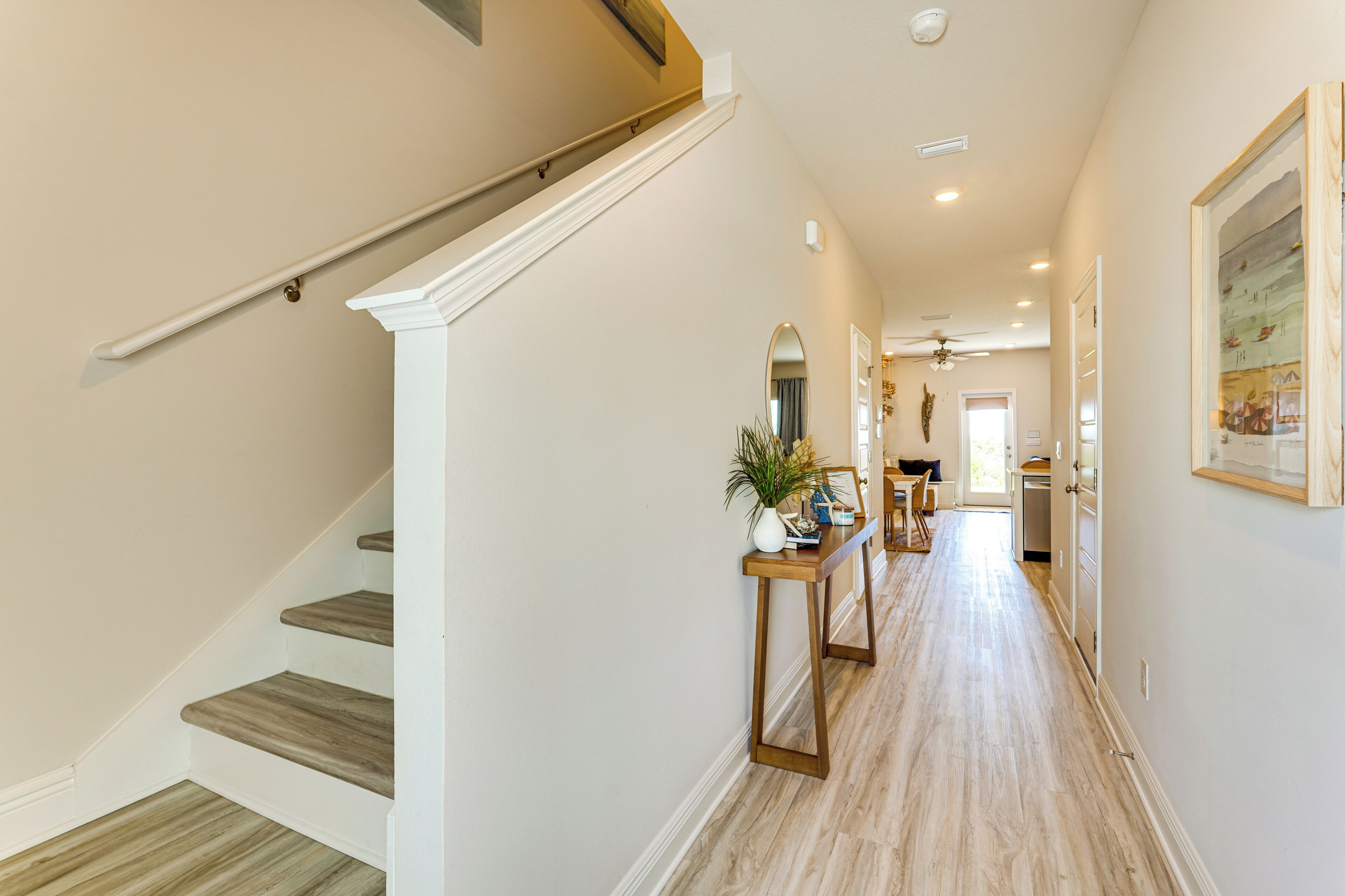 Un couloir avec un escalier en bois, une console en bois ornée d’une plante et une porte donnant sur un espace cuisine.