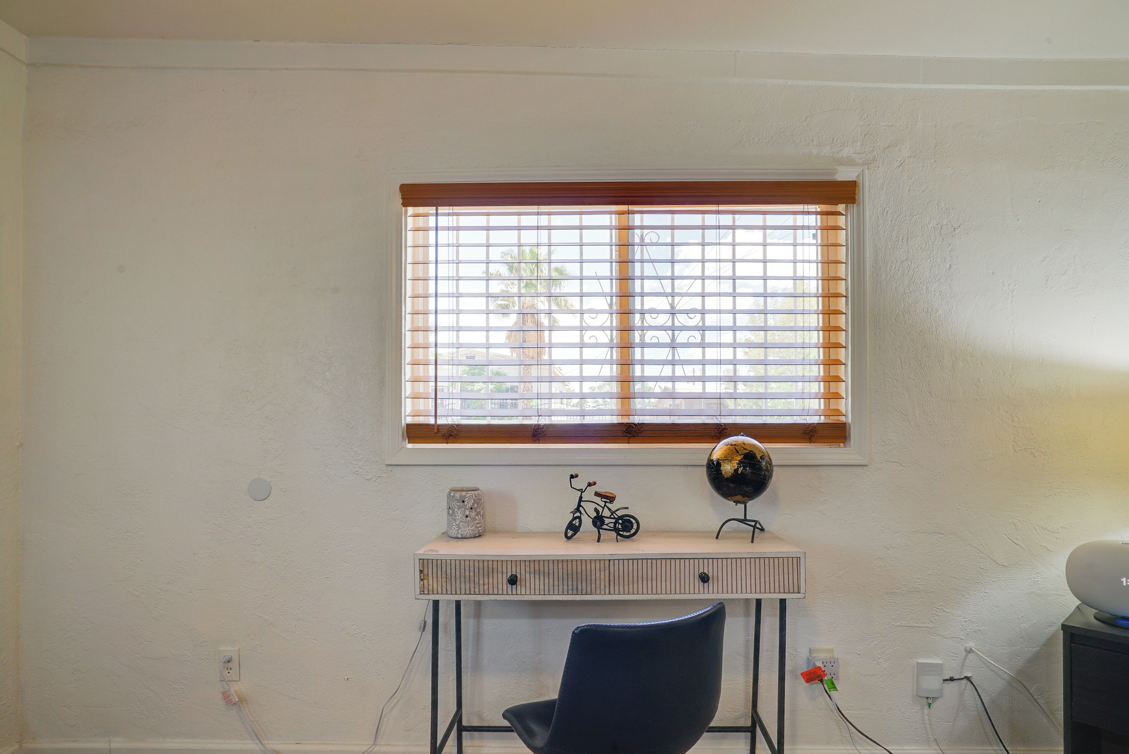 A room with a white wall, a window with blinds, a desk with a globe, a bicycle model, and a chair.
