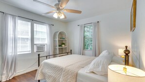A bedroom with a bed, a ceiling fan, a window with curtains, a bedside lamp, and a wall-mounted shelf with a plant. - Heart of OKC Plaza District: Duplex Near Shops! (Oklahoma City)