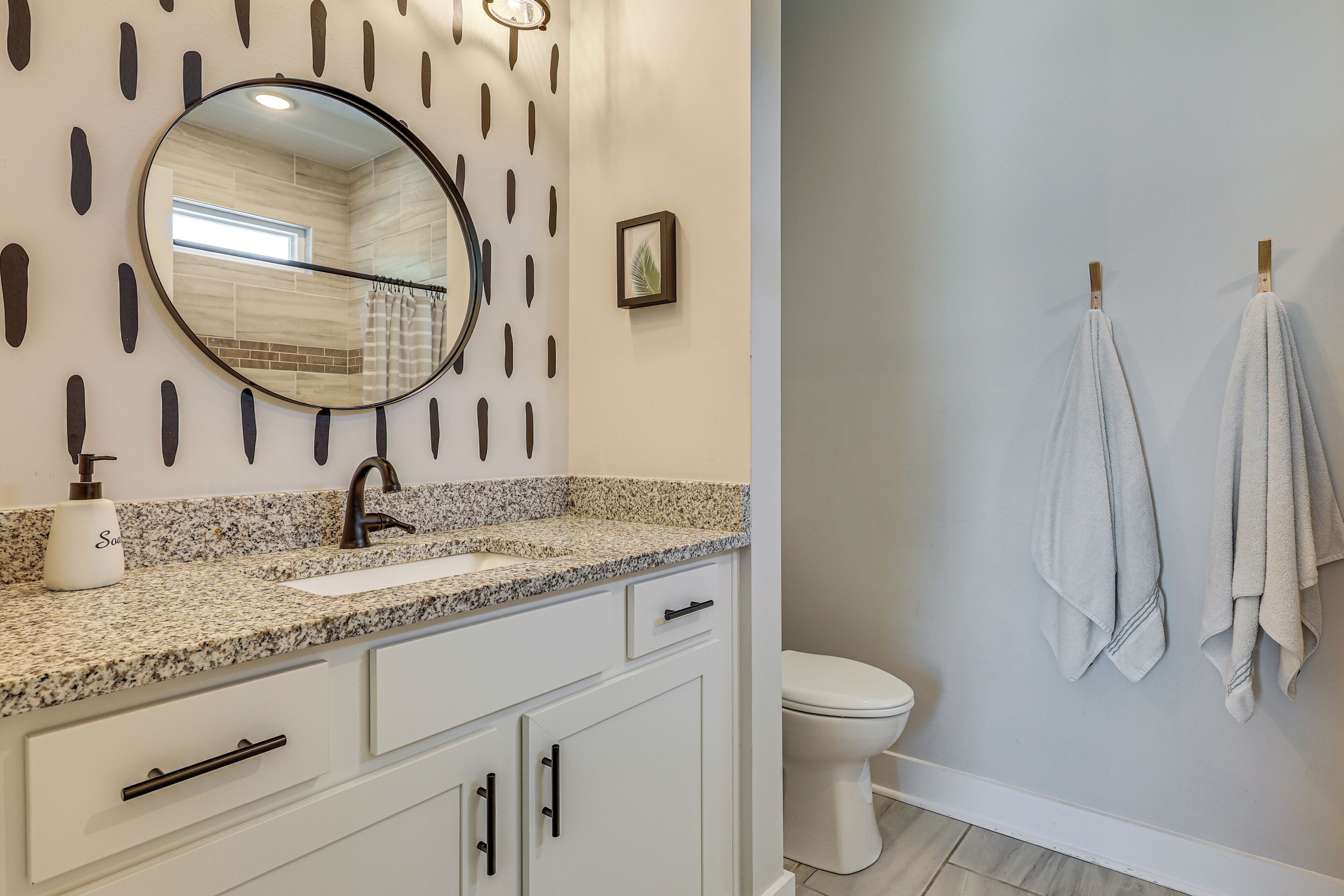 A bathroom with a granite countertop, white cabinets, a round mirror, a toilet, and towels hanging on a towel rack.