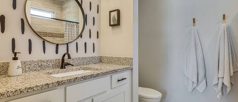 A bathroom with a granite countertop, white cabinets, a round mirror, a toilet, and towels hanging on a towel rack.