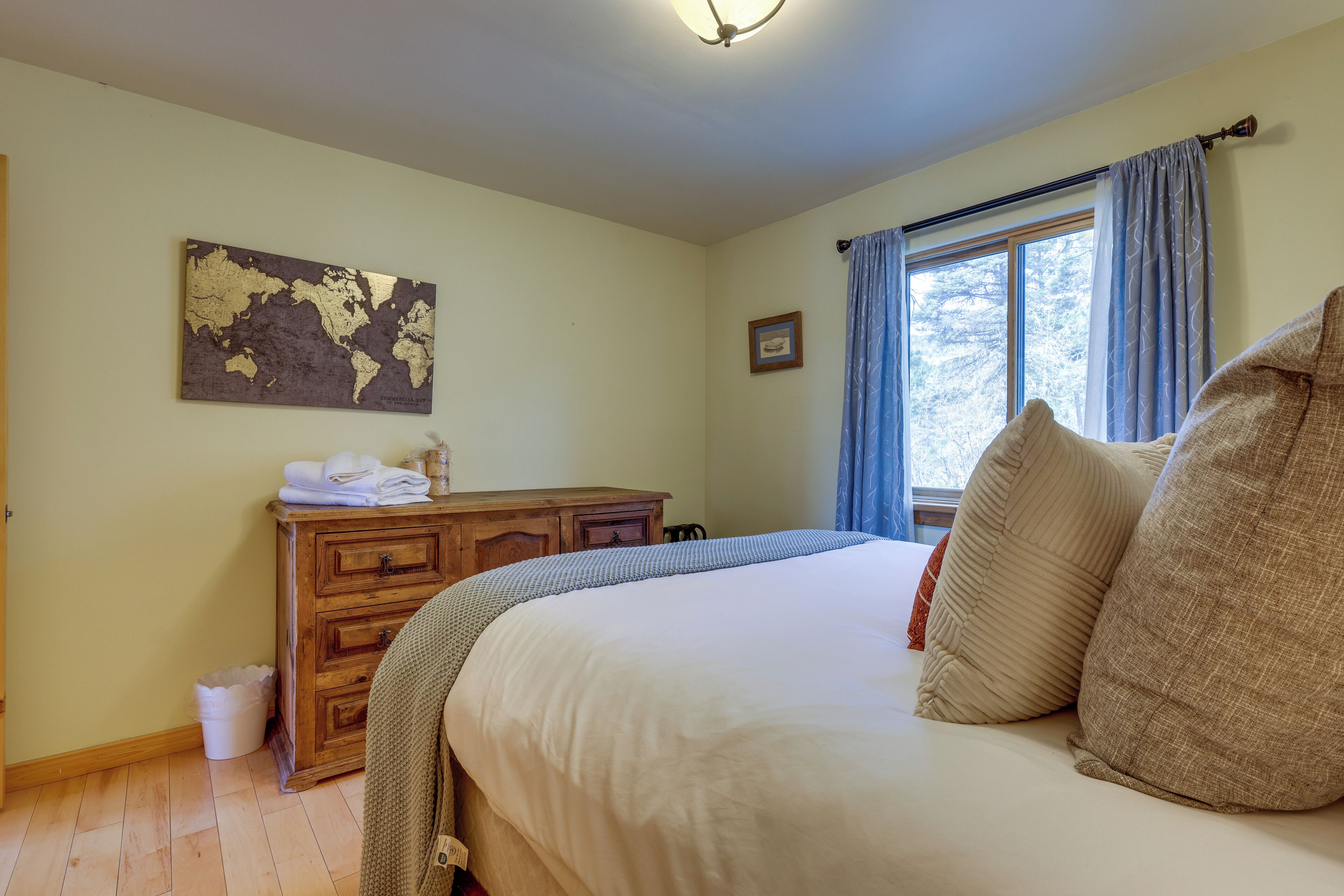 Une chambre à coucher avec un lit, une commode en bois, une carte du monde accrochée au mur, une fenêtre avec des rideaux et un petit tableau encadré.