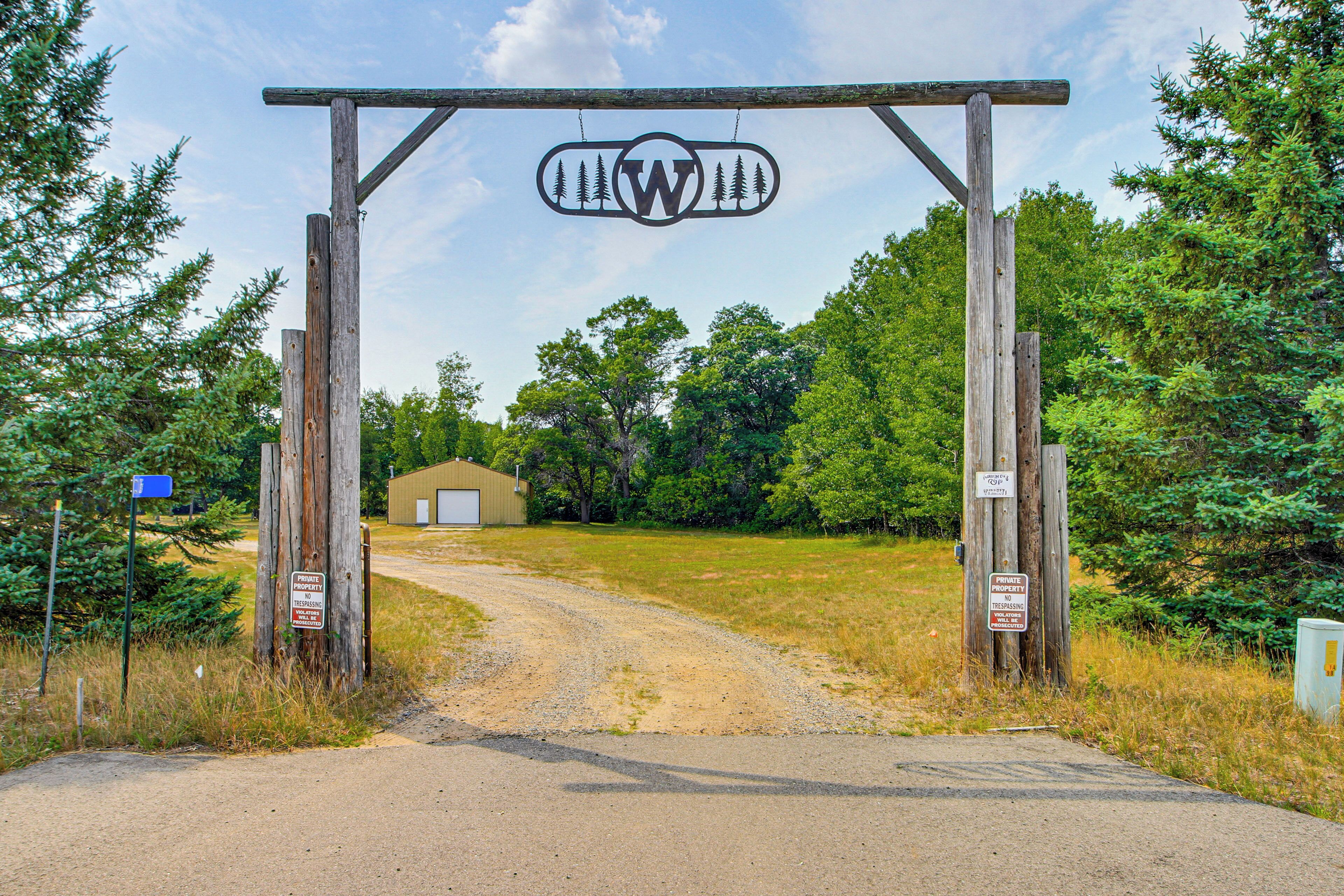 A wooden archway with a logo, a gravel path, and a building in the background.