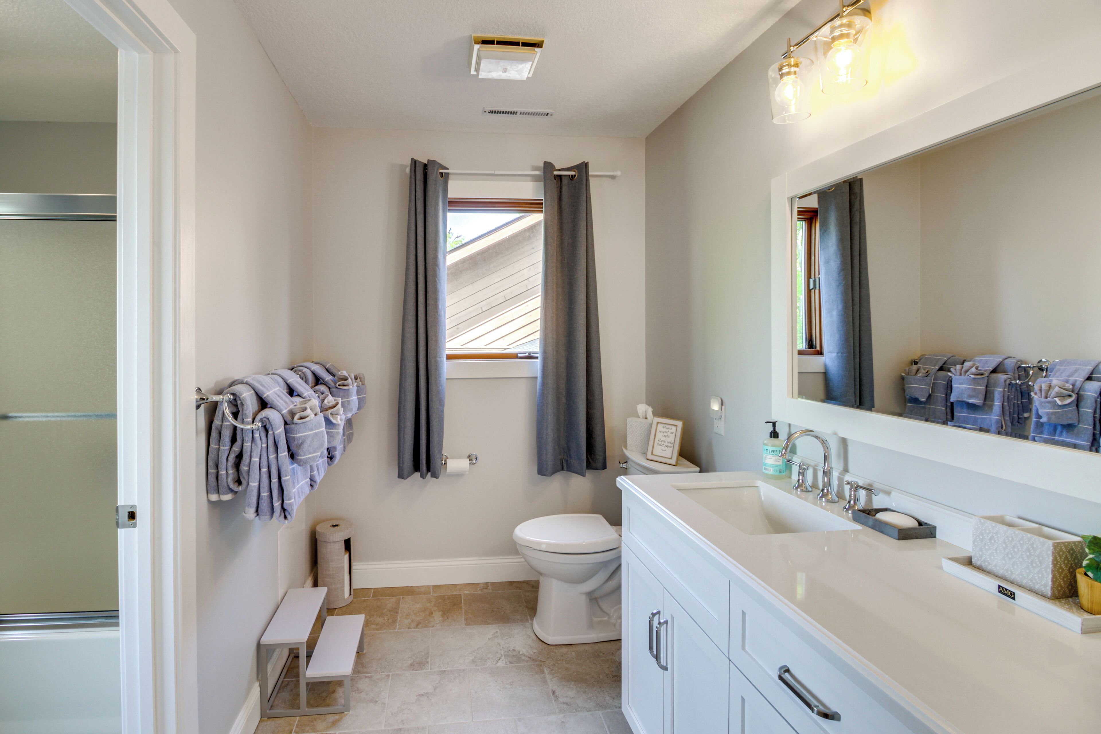 Ein Badezimmer mit Toilette, Waschbecken und Spiegel. Handtuchhalter mit Handtüchern, Duschvorhang und einer Sitzbank.