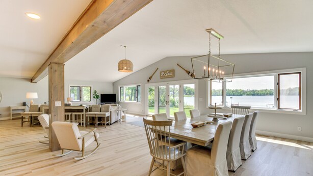 A spacious dining area with a large wooden table and chairs, a television, and a view of a lake through large windows.