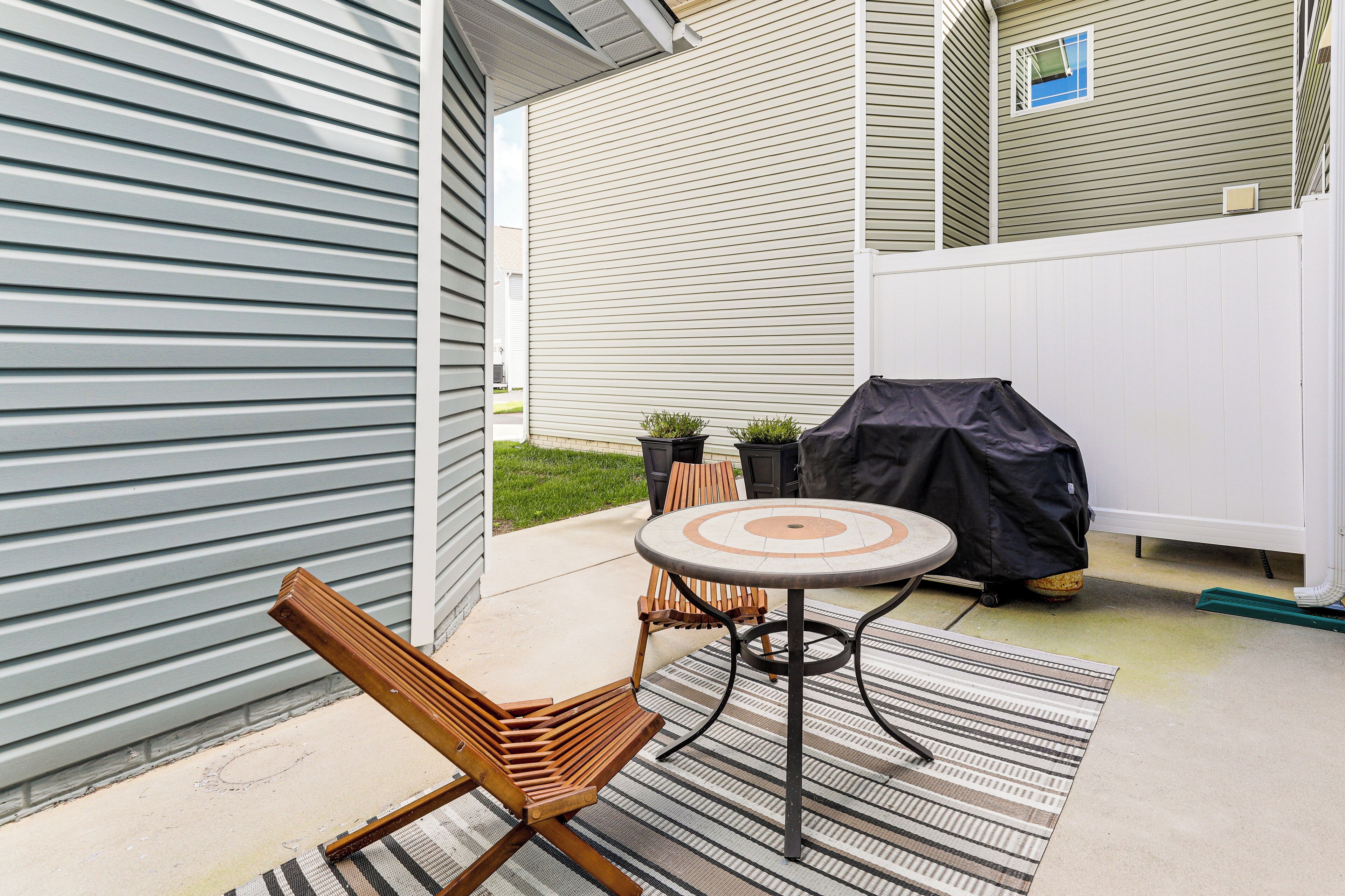 An outdoor patio with a round table, two wooden chairs, a striped rug, and a barbecue grill.