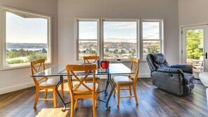 Un coin repas avec une table en verre et des chaises en bois, de grandes fenêtres offrant une vue sur les collines, et un fauteuil inclinable en cuir.
