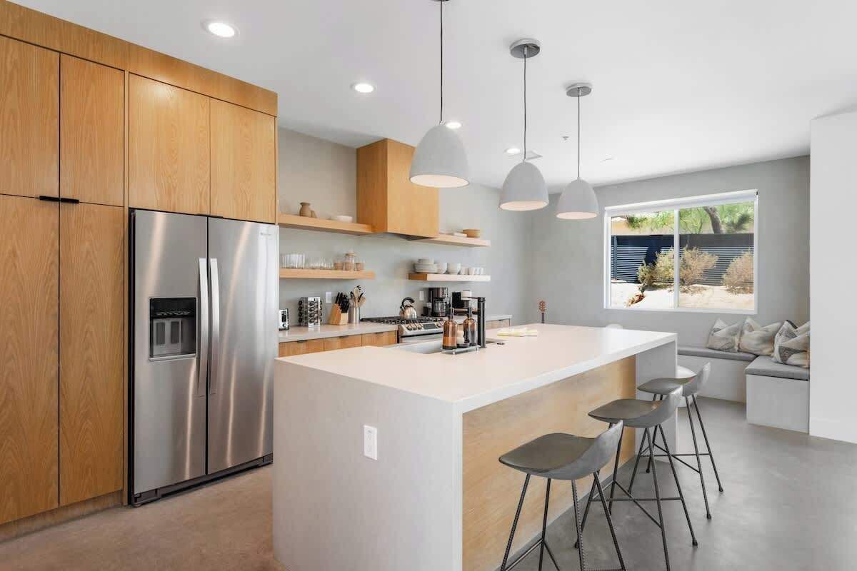 Una cocina moderna con un refrigerador de acero inoxidable, gabinetes de madera, una isla central con taburetes de bar, iluminación colgante y vistas al exterior a través de un amplio ventanal.