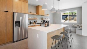 A modern kitchen with a stainless steel refrigerator, wooden cabinets, a central island with bar stools, pendant lighting, and a view of the outdoors through a large window. - Aria • Fully-Fenced Pool & Hot Tub, Luxury Design (Yucca Valley)