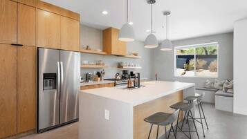 Una cocina moderna con un refrigerador de acero inoxidable, gabinetes de madera, una isla central con taburetes de bar, iluminación colgante y vistas al exterior a través de un amplio ventanal.