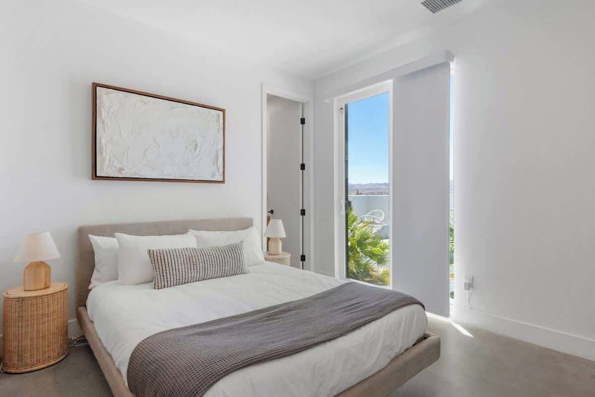Un dormitorio moderno con una cama grande, vistas a zonas verdes a través de una puerta abierta y un cuadro abstracto enmarcado en la pared.